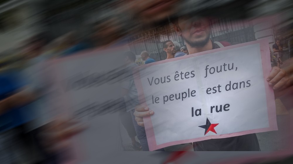 Ein Algerier hält ein Plakat mit schwarz-rotem Stern und der Aufschrift "Vous êtes foutu, le peuble est dans la rue" in die Kamera