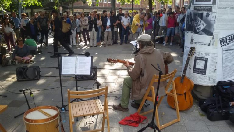 EIn Mann sitzt nebem dem Denkmal von Paulf Wulf und spielt auf einer Gitarre. Im Hintergrund stehen Zuhörer*innen